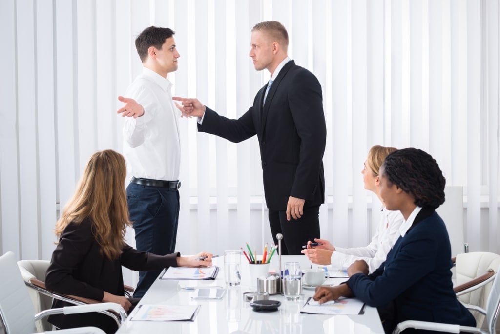 managing-high-performce-bullies Two men standing. One poking the other in the chest while three women sit at table.